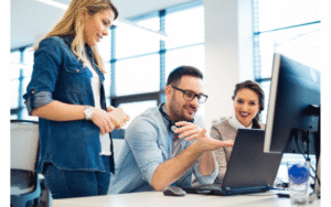 Two women and a man in office looking at a computer, with the man talking about what he is seeing.